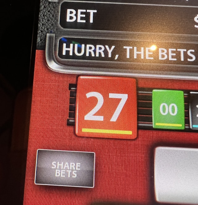 A digital display screen showing a casino betting interface, with text prompting to hurry bets and a highlighted number 27 in red. There are options for betting amounts and a 'share bets' button.